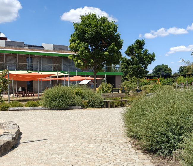 Blick auf den Garten der Kindertagesstätte Katharina von Bora mit gepflastertem Weg, Bäumen, Sträuchern und orangefarbenen Sonnensegeln.